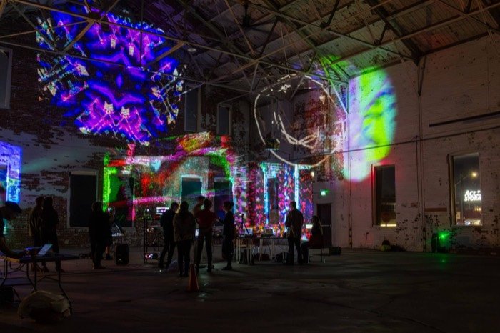 Kaleidoscopic purple and pink projections covering the walls and ceiling trusses as attendees mingle at an OPTICKS Open Lab