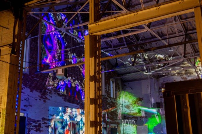 Layered purple and green projections seen through the steel roof trusses of the warehouse space