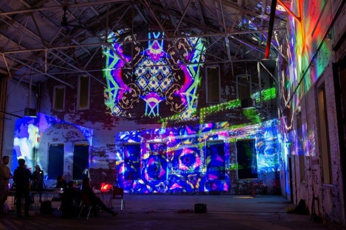 Kaleidoscopic blue and violet patterns projected across the back wall and gable of the warehouse